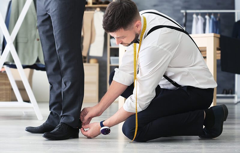 A crouching tailor adjusts and pins the hem of a client’s dress pants.