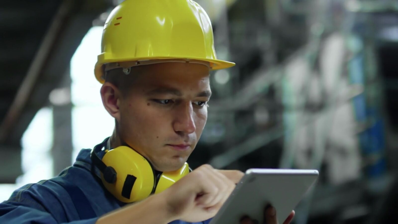 A young plant worker in a hard hat, ear defenders round his neck, works through something on a tablet.
