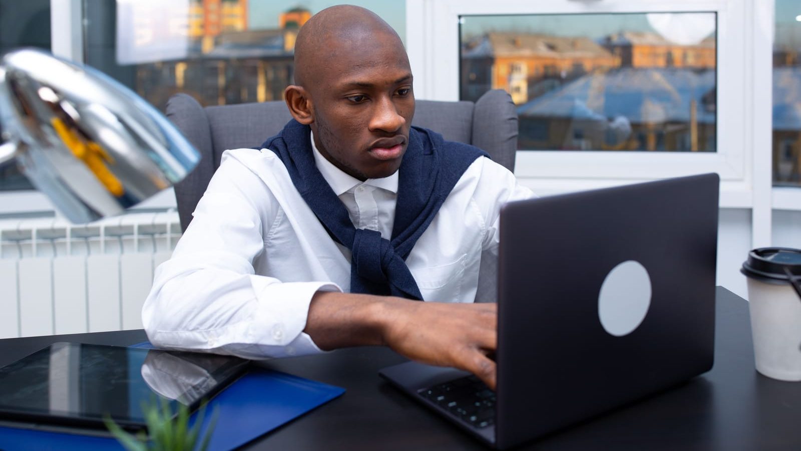 A man in a white shirt works through something on his laptop at a desk, concentrating.