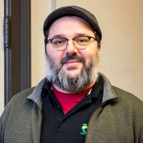 Man with a slight smile and a black and gray beard, wearing glasses and a dark hat.