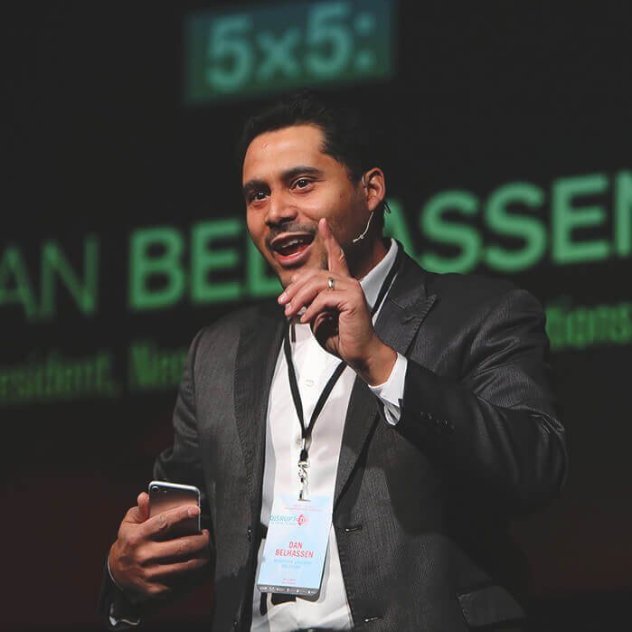 Dan Belhassen speaking on stage with a headset microphone, one finger raised to make a point