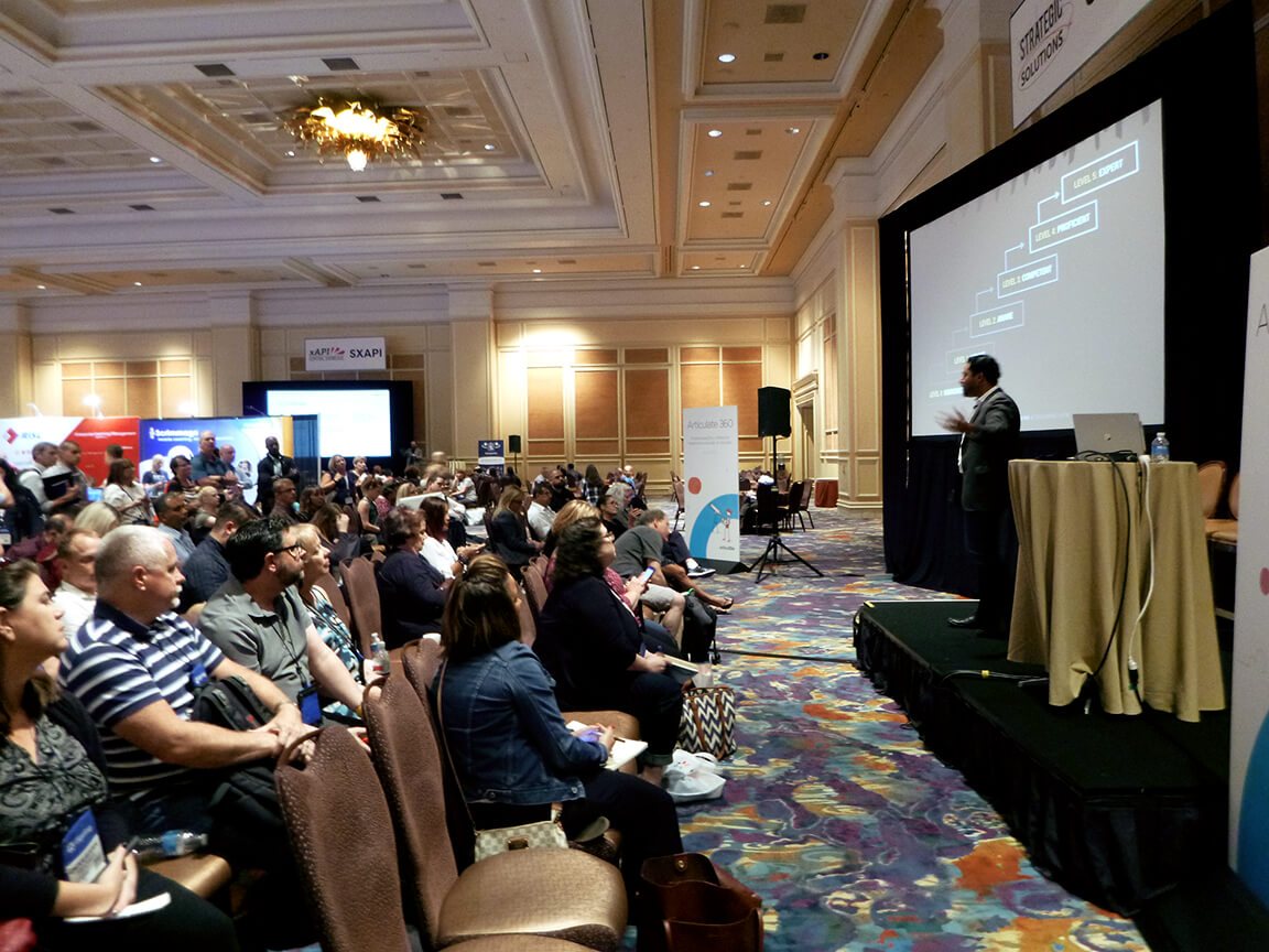 Dan Belhassen on a low stage at DevLearn, presenting to a seated audience beside a slide of mastery levels